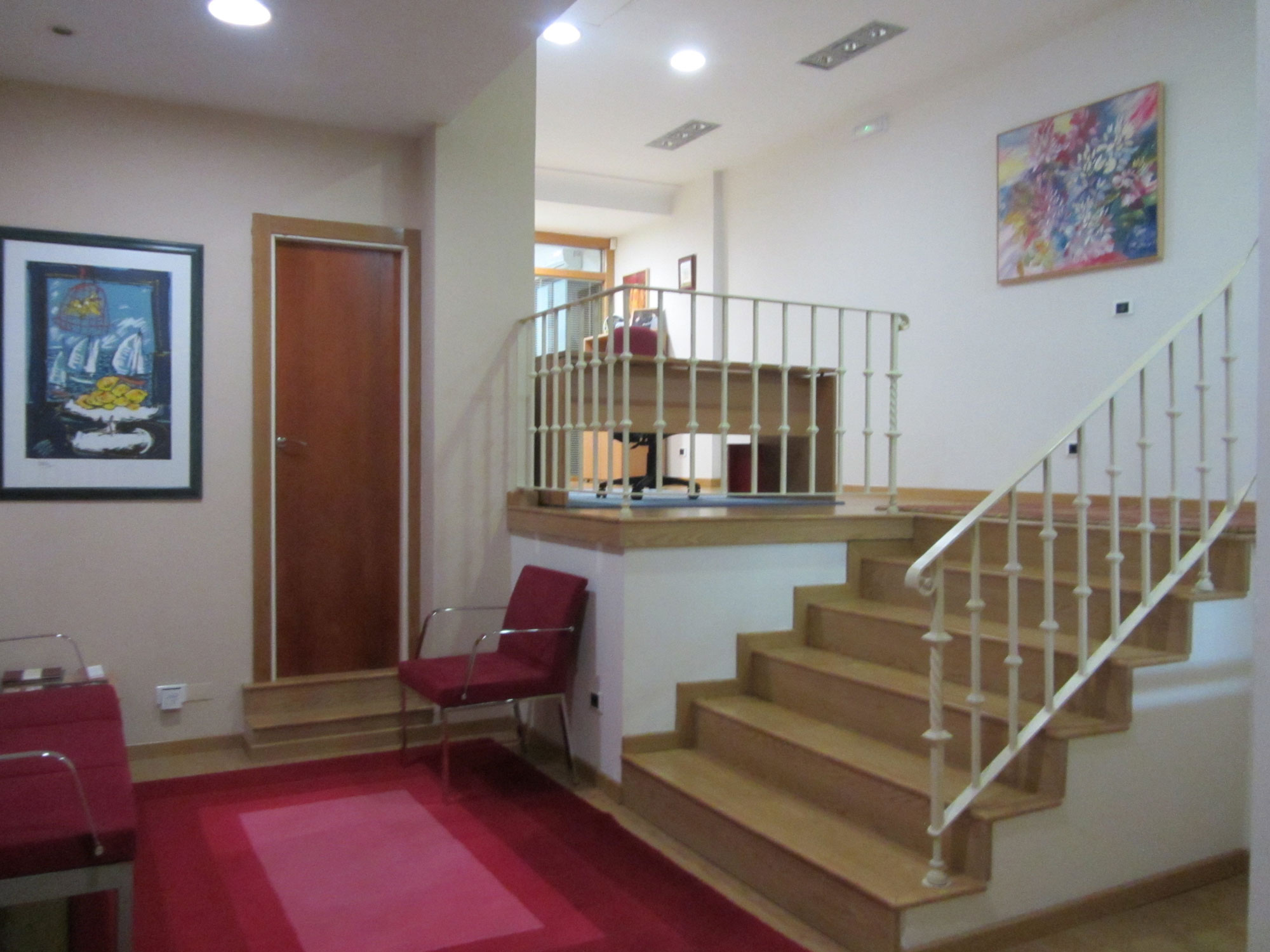 planta baja de las oficinas de ESES Galicia, se ven las escaleras para subir a la parte superior, una puerta y el suelo tiene una alfombra roja, también hay un sofá, una silla y varios cuadros en la pared